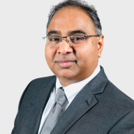 A man wearing glasses, a dark grey suit, white shirt, and grey tie poses against a plain light background, looking at the camera with a slight smile. EFESO Management Consultants